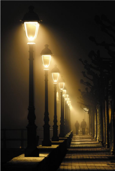 Row of illuminated street lamps along a foggy pathway with two people walking in the distance at night.